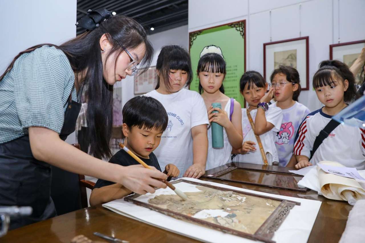 2026年7月2日，在位于山东省青岛市李沧区档案馆举办的“美在胶东 物华遗珍”李沧区百年民俗藏品展，文物修复师指导小朋友学习体验文物修复技艺。张鹰 摄 (1)